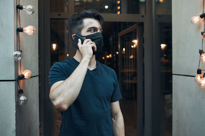 Young man standing in front of restaurant talking on phone using smartphone wearing the face mask