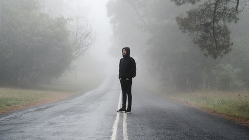 Full length of man standing on country road in forest during foggy weather