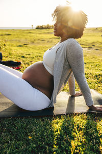 Side view of pregnant woman sitting on exercise mat over grass during training