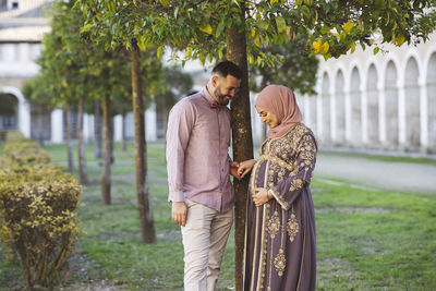 Happy husband looking at wife touching abdomen by tree in park