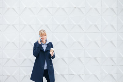 Full length of a woman using phone while standing against wall