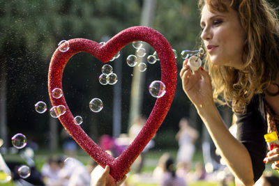 Woman blowing bubbles on red heart shape while standing outdoors