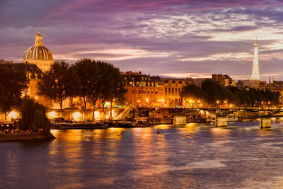 Illuminated buildings by river against sky at dusk