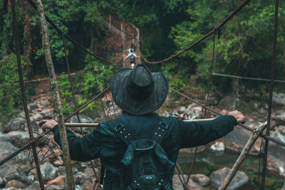 Rear view of man standing on rope bridge over stream in forest