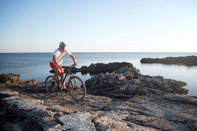 Man riding bicycle on rock by sea against clear sky