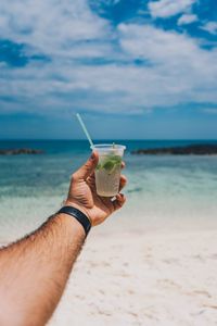 Cropped image of hand holding drink at beach against sky