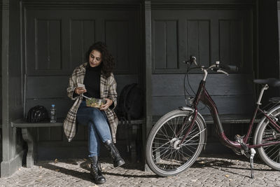 Woman with bicycle having lunch and using smartphone on station platform, berlin, germany