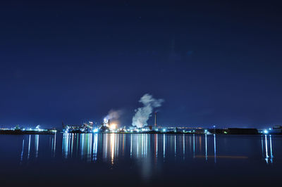 Illuminated factory by sea against sky at night