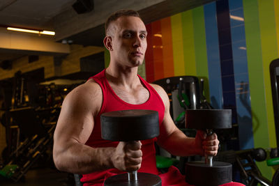 Young man holding dumbbells sitting at gym