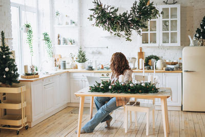 Attractive smiling woman with curly hair in plaid shirt with laptop at bright kitchen at home