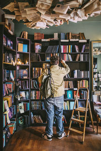 Rear view of young man with backpack choosing books from shelf in bookstore