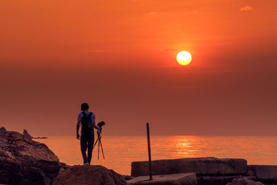 Man with tripod walking at beach against sky during sunset