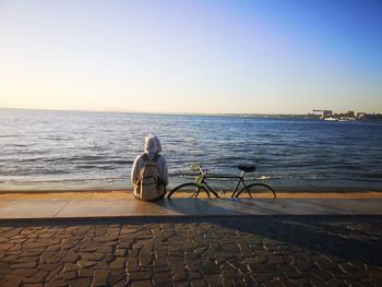 Rear view of man sitting on beach against clear sky during sunset