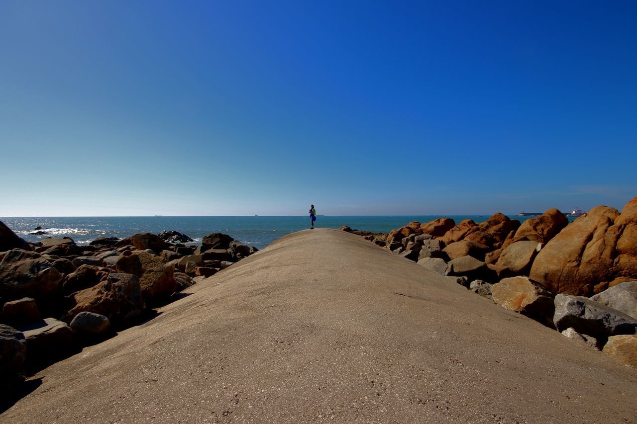 sea, water, blue, horizon over water, clear sky, beauty in nature, copy space, rock - object, tranquil scene, nature, scenics, groyne, tranquility, day, rear view, outdoors, real people, sky, one person, pebble beach, people