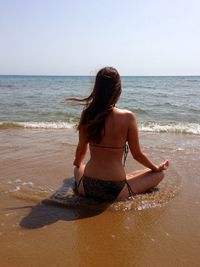 Rear view of woman in bikini meditating while sitting at beach against sky