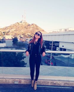 Full length of young woman wearing sunglasses standing on footbridge against clear sky