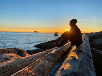Silhouette of a man sitting on logs looking out at the water during a sunset