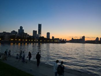 People on river by buildings against sky during sunset
