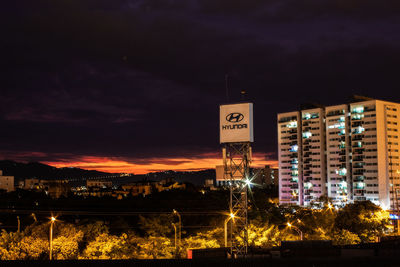 Illuminated information sign on road by buildings against sky at night