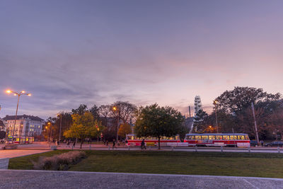 Street by illuminated city against sky at sunset