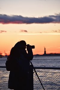 Silhouette woman photographing by sea against sky during sunset