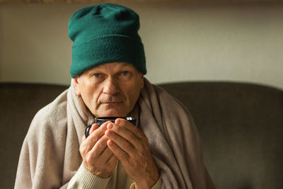 Senior man in green cap wrapped in blanket and drinking hot cup of tea, feeling cold at home.