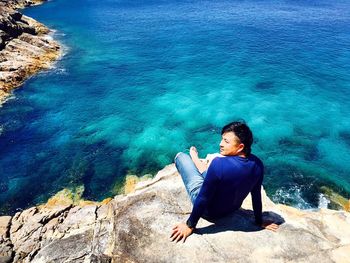 High angle view of man sitting on cliff against blue sea