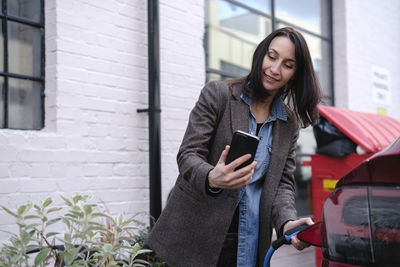 Smiling woman using smart phone and charging car through electric plug at station