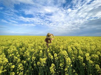 Rear view of woman wearing dress and hat in a field of canola flowers on a cloudy day