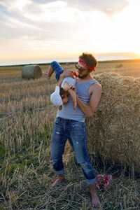 Father and son in t-shirts standing next to a haystack on a sloping field during sunset