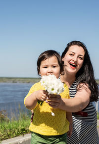 Portrait of smiling mother and daughter holding flower against sky