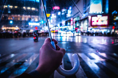 Cropped hand holding umbrella in illuminated city at night during rainy season