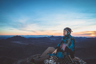 Woman looking away while sitting on mountain against sky during sunset