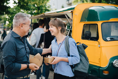 Happy male and female friends talking while standing with food box against commercial land vehicle