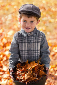 Portrait of smiling boy holding autumn leaves in bucket outdoors