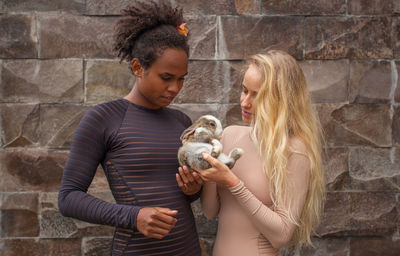 Female friends holding rabbit while standing against wall