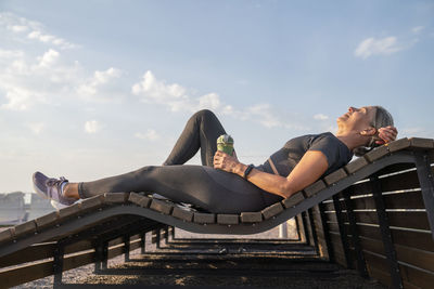 Woman holding water bottle relaxing on wooden bench on sunny day