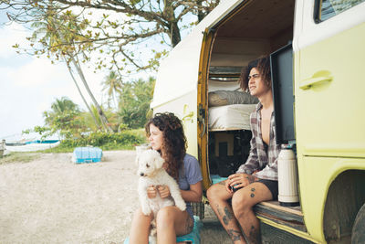Couple of travelers in casual wear with dog sitting in opened caravan car during road trip together on summer day