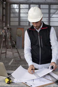 Male architect analyzing blueprint on table while standing inside construction site