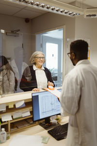 Smiling senior woman discussing with male receptionist through transparent shield in clinic