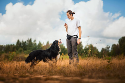 Man training dog on grassy land against cloudy sky