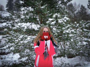 Portait of a woman in winter clothes wearing red gloves with hearts standing near a snowy pine tree