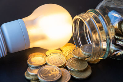 Fluorescent light bulb turned on, and jar full of coins next to it. increased costs. energy tariff.