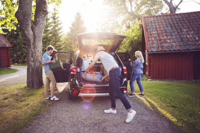 Full length of friends loading luggage into car on road during sunny day