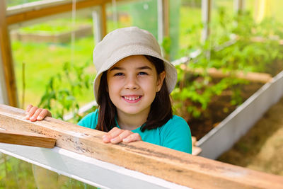 Portrait of a happy little girl in a white panama, looks out of the window of a wooden  greenhouse