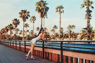 Full length portrait of young woman leaning on railing against palm trees