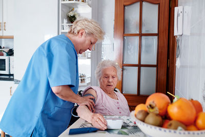Caring nurse with professional tonometer measuring blood pressure to aged woman sitting at table with food in kitchen at home