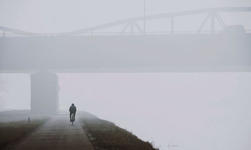 Rear view of person riding bicycle by lake on road during foggy weather