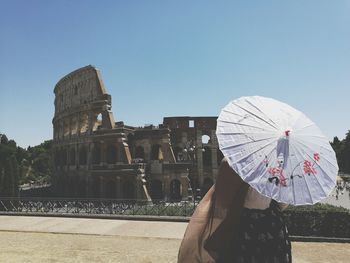 Rear view of woman with umbrella standing by coliseum against clear blue sky during sunny day
