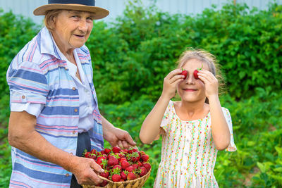 Smiling girl covering eyes with strawberries standing by grandmother